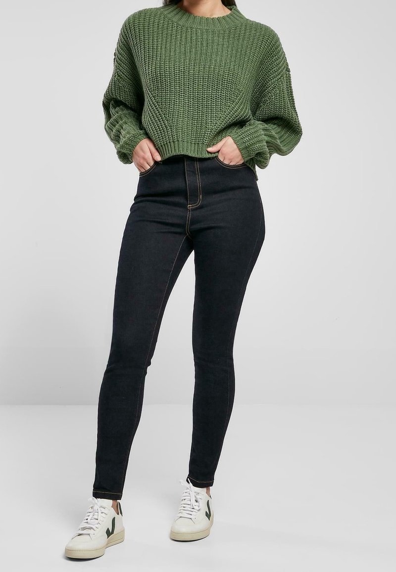 Pull en maille verte avec une texture côtelée, associé à un jean skinny noir taille haute. Les chaussures sont des baskets blanches avec des accents verts.
