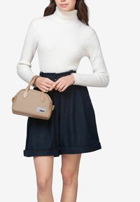 Tan håndtaske med afrundede kanter og dobbelt håndtag, der har en rektangulær bund og et synligt mærkat. Modellen bærer en hvid ribbet turtleneck og marineblå shorts.