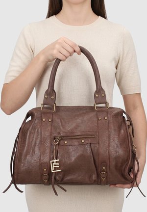 Donna che tiene una grande borsa in pelle marrone con cerniera dorata e charm decorativo, indossa una maglietta crema a maniche corte.