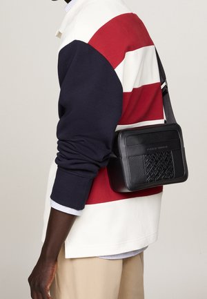 Borsa a tracolla nera rettangolare con fronte texturizzato e lati lisci. Cintura regolabile; persona che indossa un maglione a righe crema e blu navy.