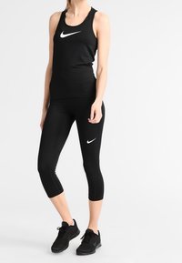 Débardeur noir et leggings capri fabriqués dans un matériau extensible, ornés de logos Nike swoosh blancs. Portés avec des chaussures de sport noires.