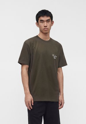 Młody mężczyzna z krótkimi ciemnymi włosami, ubrany w oliwkowo zielony T-shirt z małym białym napisem na lewej piersi, stojący na jednolitym jasnym tle.