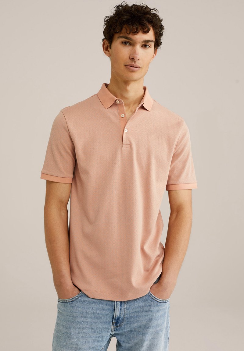 Polo cu mânecă scurtă de culoare roz pastel, cu un model geometric subtil, guler ribbed și închidere cu două butoane. Purtați-l cu blugi albaștri deschiși.