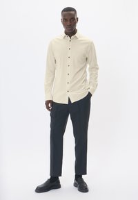 Hosszú ujjú krémszínű ing sötét gombokkal, sötét nadrággal és fekete félcipővel párosítva, egy fehér háttér előtt állva.