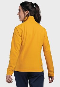 Gelbe Fleece-Jacke mit hohem Kragen, verfügt über eine flache Nahtkonstruktion und eine glatte Textur, von hinten mit einem Pferdeschwanzfrisur zu sehen.