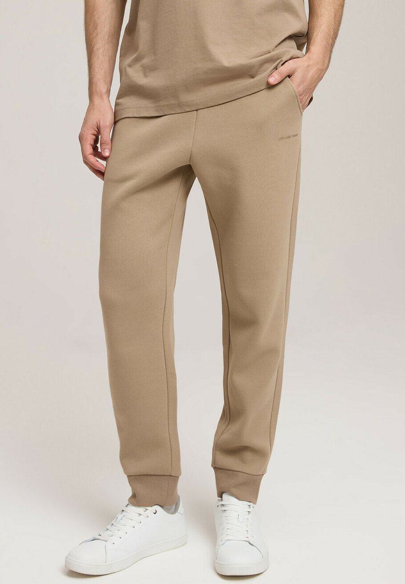 Beżowe dresy wykonane z gładkiej mieszanki bawełny, z tapered designem, kieszeniami po bokach i ściągaczami, w połączeniu z białymi sneakersami.