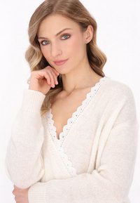 Biały sweter dzianinowy z falbankowym koronkowym wykończeniem, dekoltem w serek i miękką fakturą. Posiada bufiaste rękawy oraz luźny krój.