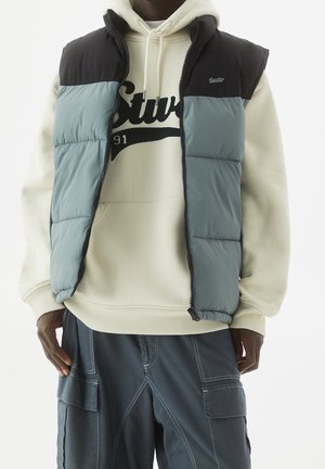Homme portant une doudoune noire et grise par-dessus un sweat à capuche crème avec texte noir, associée à un pantalon cargo gris foncé avec des poches.