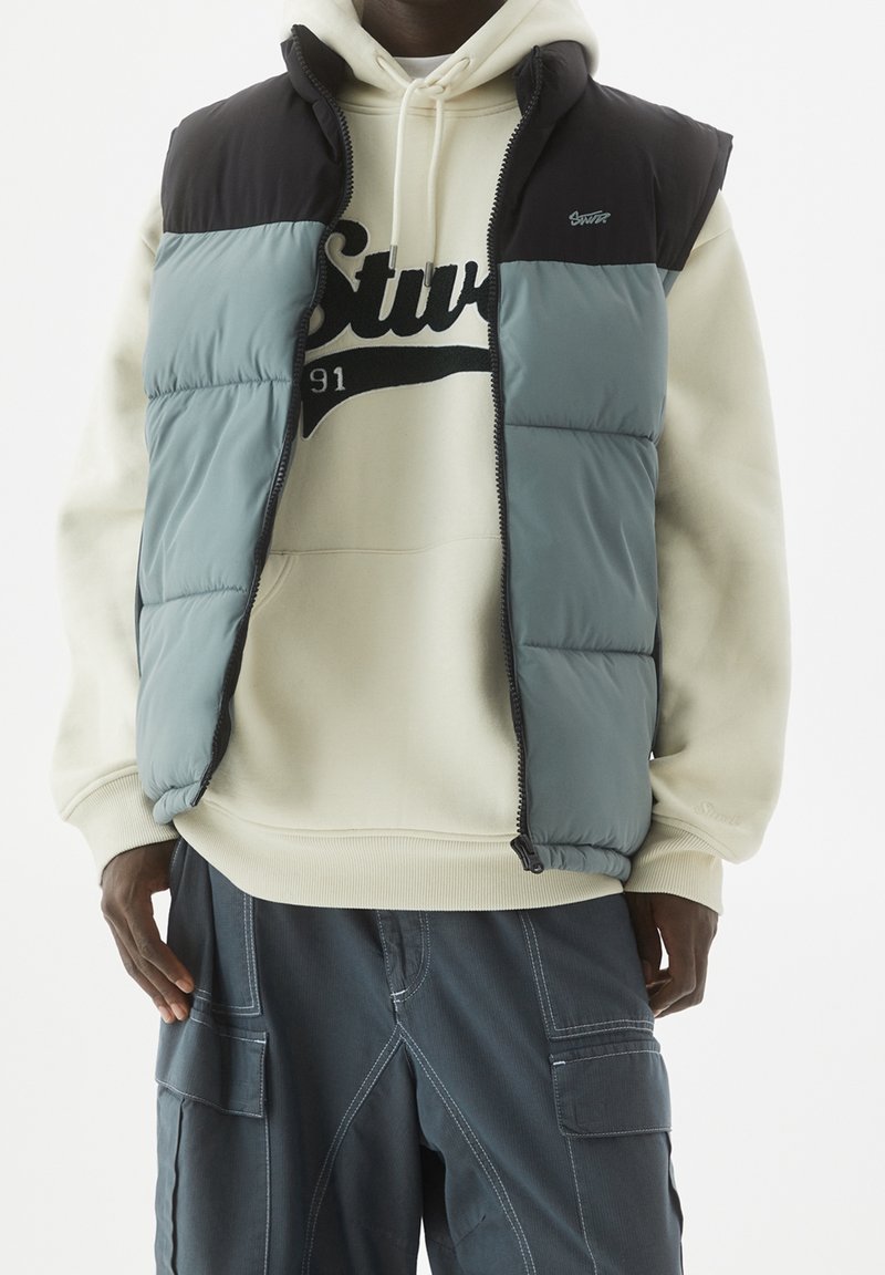 Homme portant une doudoune noire et grise par-dessus un sweat à capuche crème avec texte noir, associée à un pantalon cargo gris foncé avec des poches.