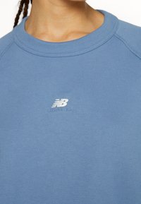 Personne portant un sweat-shirt New Balance bleu clair avec une coiffure tressée visible sur le côté gauche, gros plan du cou à la poitrine.