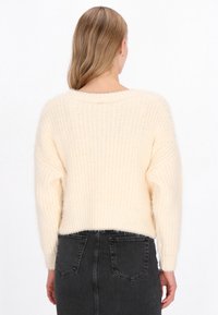 Krém színű, bolyhos kötött pulóver, laza szabással és bordás textúrával, széles nyakkivágással és beszűkített ujjaival. Fekete szoknyával viselve.