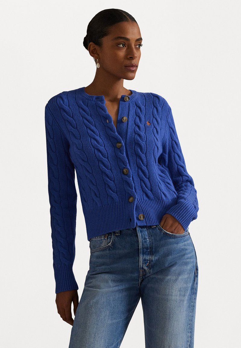 Blauwe kabelgebreide cardigan met een ronde halslijn, vijf bruine knopen en een geribbelde zoom; gedragen over lichte blauwe jeans.