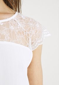 Weißes Oberteil mit enganliegendem Rippdesign und zarten Spitzenärmelchen, das komplexe florale Muster und einen gewellten Rand zeigt.