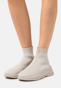 Béžové kotníkové tenisky se strukturním pleteným svrškem. Mají měkký polstrovaný límec a robustní gumovou podrážku s logem značky v zlaté barvě.