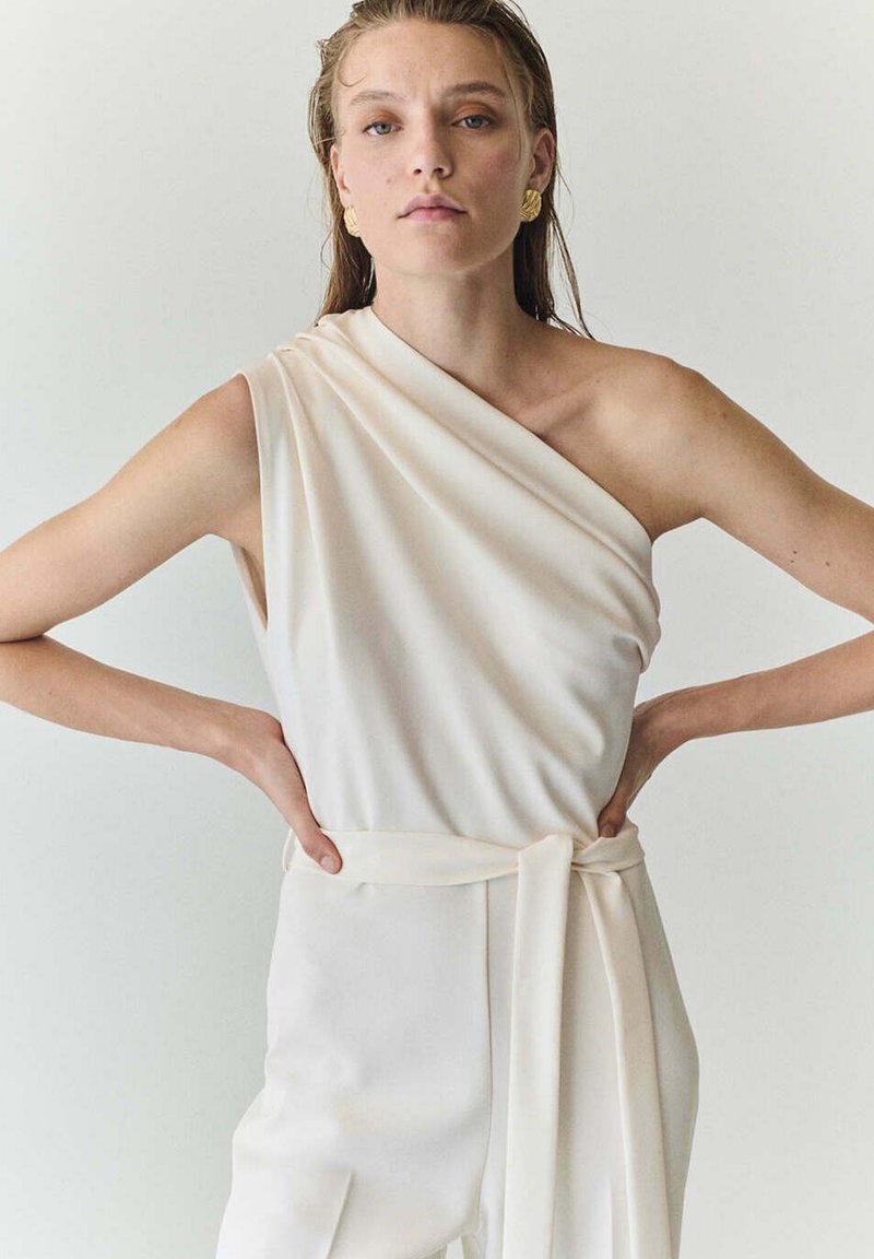 Femme portant une robe écrue à une épaule avec une ceinture nouée à la taille, debout les mains sur les hanches sur un fond uni.