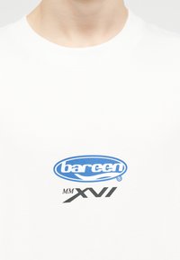 Personne portant une chemise blanche avec un logo ovale bleu indiquant "bareen" et un texte noir "MM XVI" sous le logo sur la poitrine.