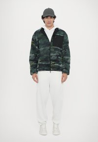 Fleece jacket in green camouflage pattern with a black zippered pocket, worn over a white hoodie, paired with white sweatpants and sneakers.