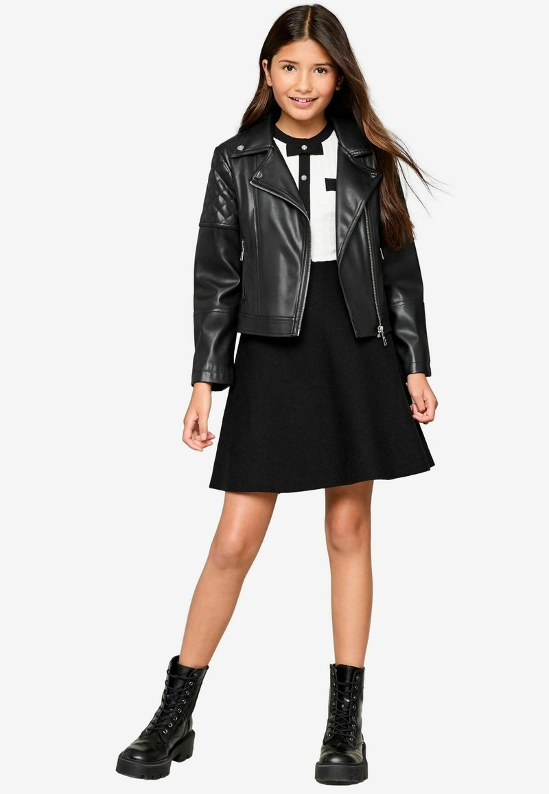 Zwarte leren motorjack met creëpte schouders, witte blouse met zwarte knopen en een zwarte klokrok. Zwarte veterschoenen.