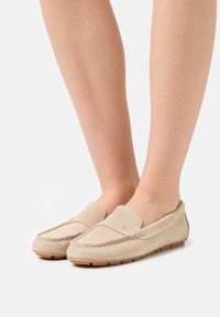 Beige ruskindsloafers med en syet detalje, flad gummisål og rund tå. Har en dekorativ flap foran.