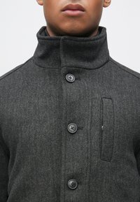 Casaco cinza escuro com padrão espinha de peixe, gola alta, três grandes botões pretos e detalhe de bolso lateral.