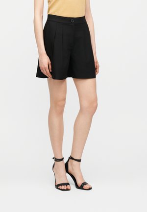 Femme portant un short plissé noir, un haut jaune et des sandales à talons hauts noires avec bride à la cheville, debout devant un fond blanc uni.