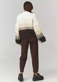 Gruby sweter pleciony w gradientowe paski w kolorze kremowym, szarym i brązowym. Posiada długie, frędzlowe rękawy. Połączony z brązowymi spodniami i czarnymi butami.