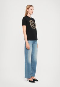 Frau im Seitenprofil, die ein schwarzes Graphic-T-Shirt, hellblaue weite Jeans und schwarze spitze High Heels vor einem weißen Hintergrund trägt.