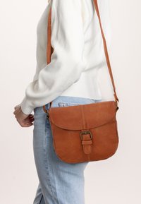 Asmuo baltu megztiniu ir šviesiai mėlynomis džinsomis, ant klubo nešantis rudą odinę per petį kabinamą krepšį su užsegimu.