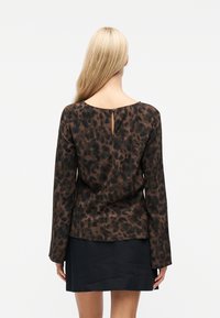 Femme aux longs cheveux blonds portant un haut à manches longues imprimé léopard marron et noir et une courte jupe noire, vue de dos sur un fond blanc.