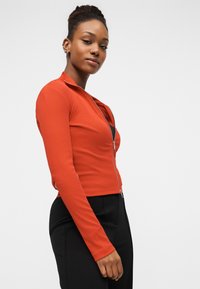 Κόκκινο στενό μπουφάν με φερμουάρ, μακριά μανίκια, ριμπ υφή και γιακά. Συνδυασμένο με μαύρο ψηλόμεσο παντελόνι.