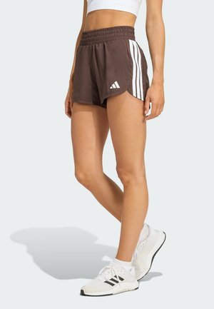 Atlet iført brune Adidas-shorts med hvide striber og logo, hvid crop top og hvide sneakers stående på lys baggrund.
