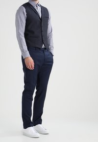 Mężczyzna w niebieskiej koszuli, czarnym kamizelce, granatowych spodniach i białych trampkach stojący na tle jasnego, jednolitego tła.