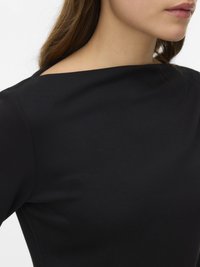 Žena se světlou pokožkou a hnědými vlasy, oblečená v černém topu s lodičkovým výstřihem, záběr zblízka oříznutý od krku po horní část hrudi.