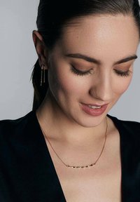 Złoty naszyjnik z skręconymi ogniwami, odbijający światło. Modelka ma na sobie proste złote kolczyki, eksponując gładką skórę i ciemny, elegancki strój.