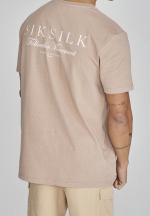 Mężczyzna ubrany w beżową koszulkę SikSilk z białym napisem "SIKSILK Follow the Movement" oraz beżowe szorty typu cargo, pokazujący tatuaż na plecach i prawej ręce.