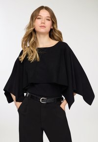 Top nero oversize in stile mantella con scollo ampio, design a strati e maniche fluenti, abbinato a pantaloni neri aderenti e una cintura.