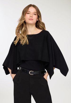 Top nero oversize in stile mantella con scollo ampio, design a strati e maniche fluenti, abbinato a pantaloni neri aderenti e una cintura.
