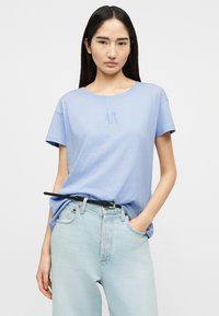 Γυναίκα με μακριά μαύρα μαλλιά, φορώντας ανοιχτό μπλε κοντομάνικο πουκάμισο και ανοιχτόχρωμα τζιν με μαύρη ζώνη, στέκεται μπροστά σε απλό φόντο.