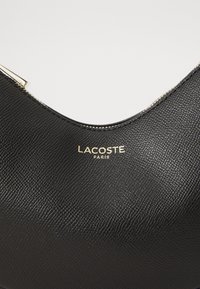 Black textured leather handbag with a curved top, featuring a gold-tone "LACOSTE PARIS" logo and a gold zipper accent.