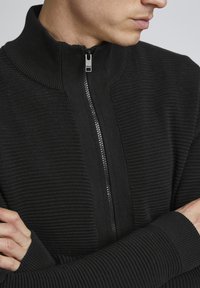 Schwarzer Reißverschluss-Pullover mit strukturiertem Rippmuster, hohem Kragen und kontrastierendem Reißverschluss, engeren Bündchen und schlankem Design.