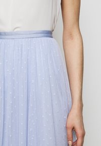 Närbild av en person som bär en vit topp och en ljusblå tyllkjol med små vita broderade prickar och ett satinliknande midjeband.