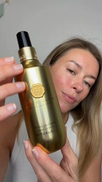 Kvinna håller en guldfärgad flaska med Rituals Hibiscus Dream hemparfym nära ansiktet mot en enfärgad bakgrund.