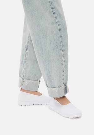 Scarpe bianche slip-on con tomaia in knit testurizzata, abbinate a jeans denim azzurri con risvolto. Le scarpe presentano una suola in gomma e un design minimalista.