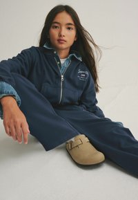 Tuta blu navy con chiusura a zip, colletto in denim e logo. Zoccoli beige con cinturino, posati su uno sfondo chiaro.