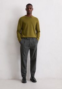 Zwarte man leunend tegen de muur in een olijfgroen trui, grijze geplooide broek en zwarte leren schoenen, met zijn handen in zijn zakken.