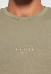 Λαδί πράσινο T-shirt κατασκευασμένο από ριπ ύφασμα, με στρογγυλή λαιμόκοψη και μικρό λογότυπο "GUESS EST. 1981" σε ανοιχτότερο μπεζ χρώμα.