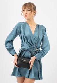Borsa a tracolla in pelle nera con una chiusura decorativa a forma di ape, forma arrotondata, manico corto e tracolla a catena removibile; indossata con un abito avvolgente di raso teal.