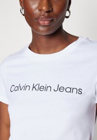 Zbliżenie osoby noszącej białą koszulkę Calvin Klein Jeans z srebrnymi kolczykami kołami, ukazujące górną część klatki piersiowej i usta.