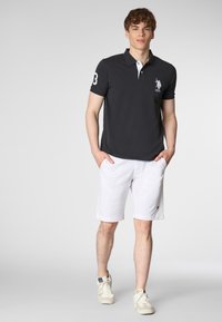 Polo grigio scuro con accenti bianchi, dotato di un logo ricamato. Abbinato a pantaloni corti bianchi e sneaker di colore chiaro.