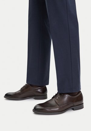 Ciemnobrązowe skórzane eleganckie buty na sznurowadła noszone z granatowymi, dopasowanymi spodniami na białym tle.